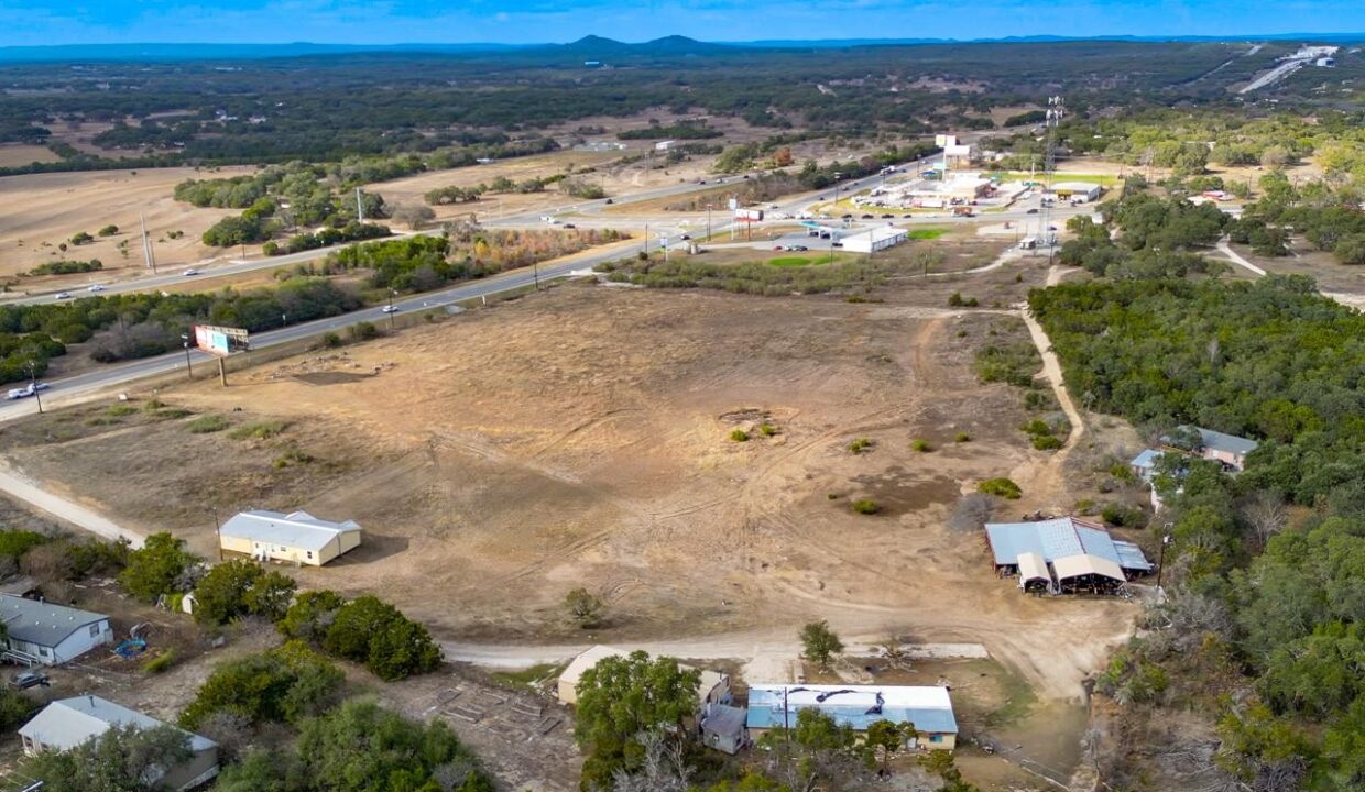 Aerial view of a rural area with open fields, scattered houses, barns, dirt roads, and a small town with various buildings in the distance under a partly cloudy sky.