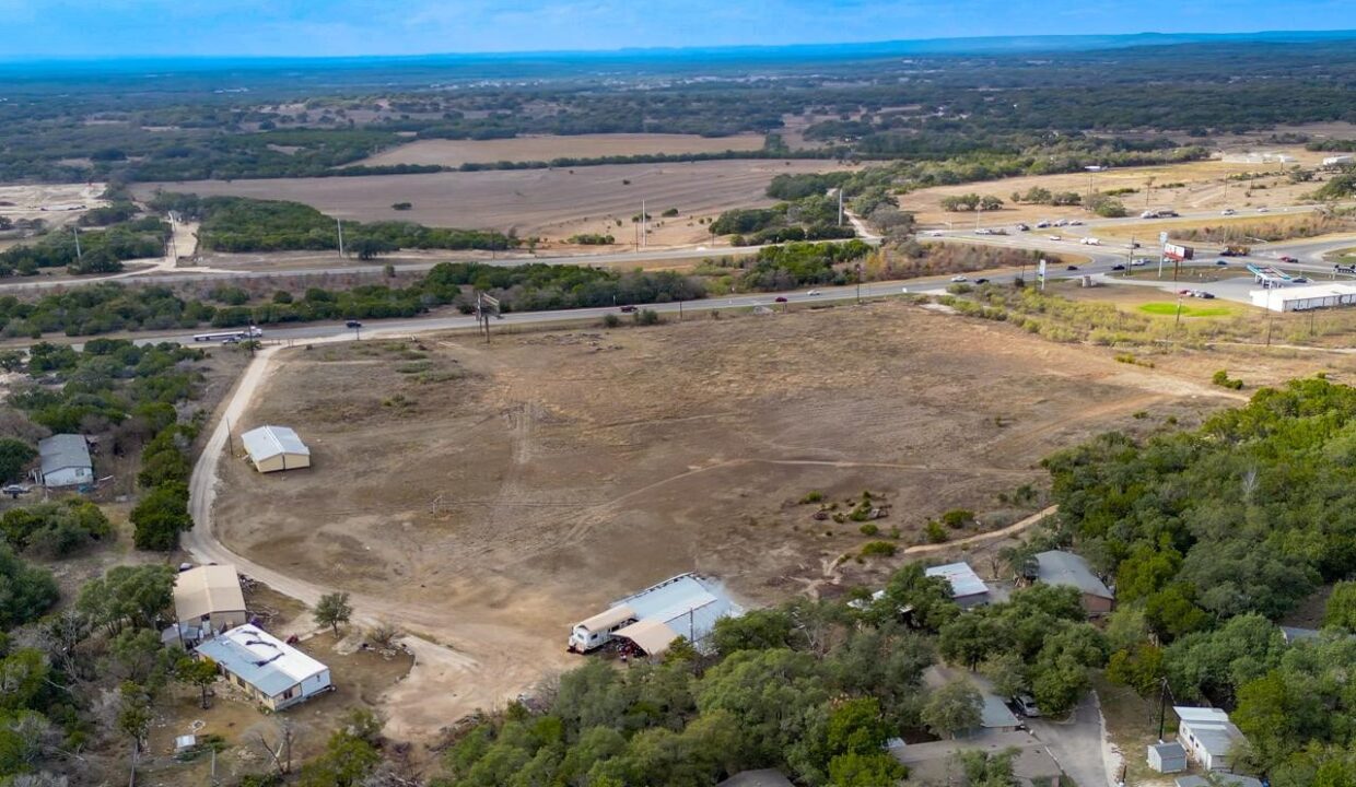 Aerial view of a large, mostly empty and dry lot bordered by trees and a few buildings, with roads and fields extending into the distance under a partly cloudy sky.