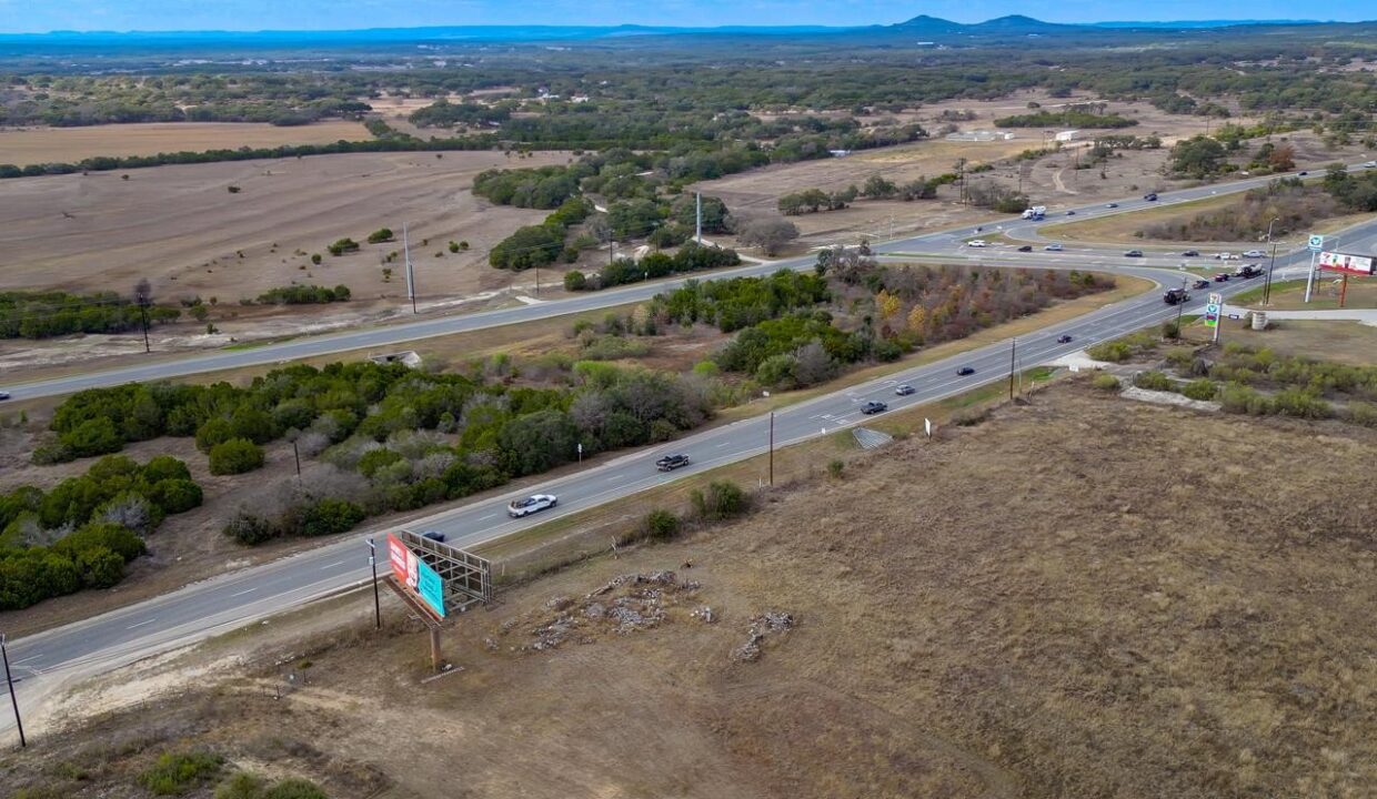 Aerial view of a rural intersection with several cars on a winding road, surrounded by dry grass fields, scattered trees, and distant hills under a blue sky with clouds. Billboards are visible near the roads.