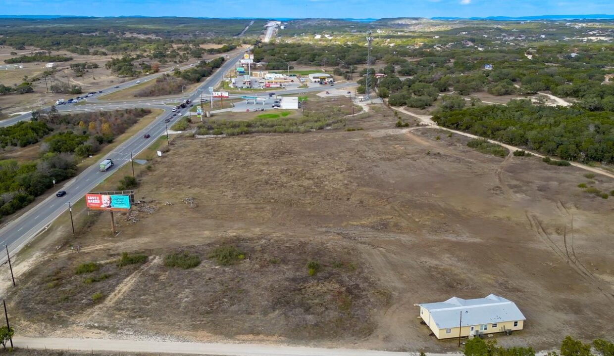 Aerial view of a rural area with a main road, cars, billboards, a cluster of commercial buildings, and a single house on a mostly empty, dry plot of land surrounded by trees and hills.