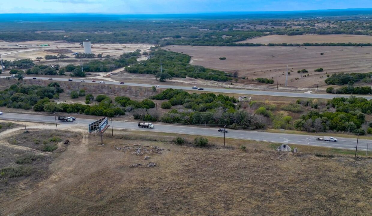 Aerial view of a rural highway with cars and trucks driving, fields and patches of trees on both sides, a billboard, and a distant water tower under a partly cloudy sky.