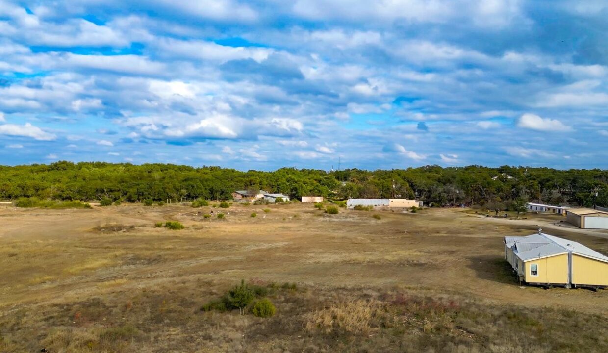 A wide, open field with dry grass stretches towards several scattered buildings and houses, bordered by dense green trees under a partly cloudy sky.