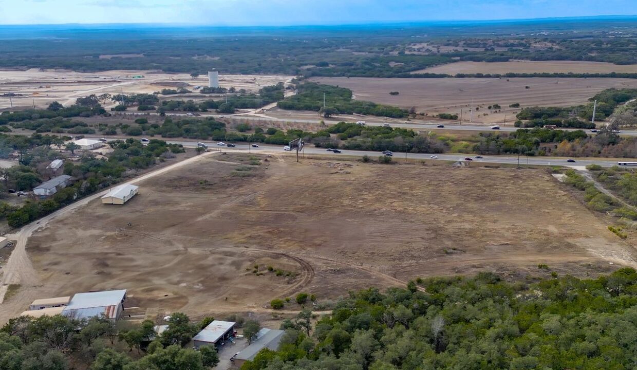 Aerial view of a large, mostly barren plot of land bordered by trees, roads, and scattered buildings, with expansive fields and distant hills under a cloudy sky in the background.