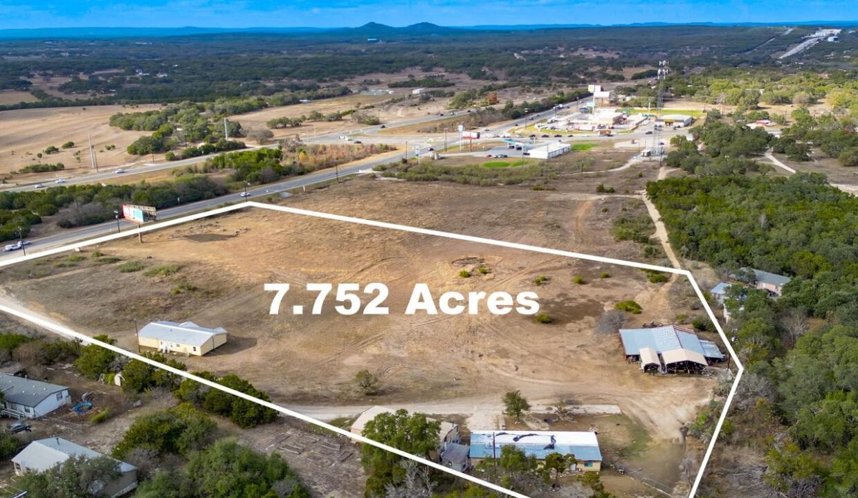 Aerial view of a 7.752-acre plot of land outlined in white, with a few buildings and trees on the property, surrounded by rural roads, scattered houses, and a distant small town.