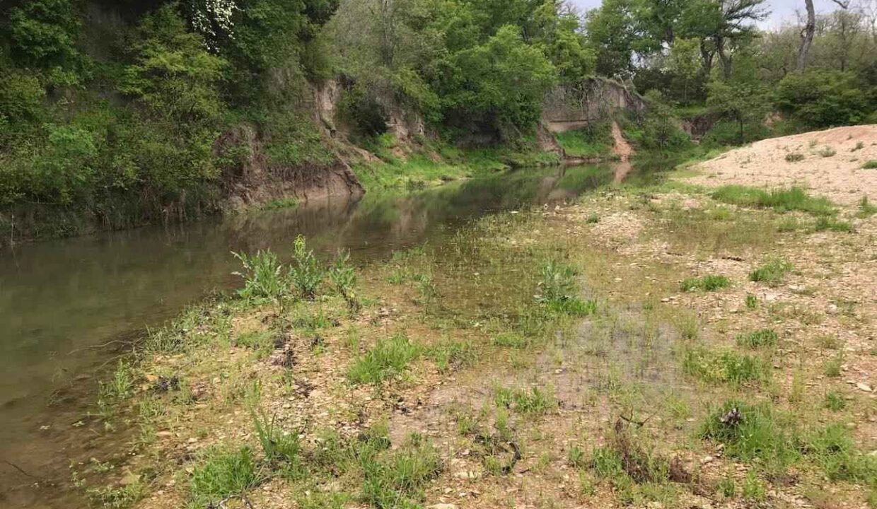 A calm river with clear water flows alongside a rocky, grassy bank. Trees with green foliage line the riverbank, and the sky appears overcast. The area looks peaceful and natural.