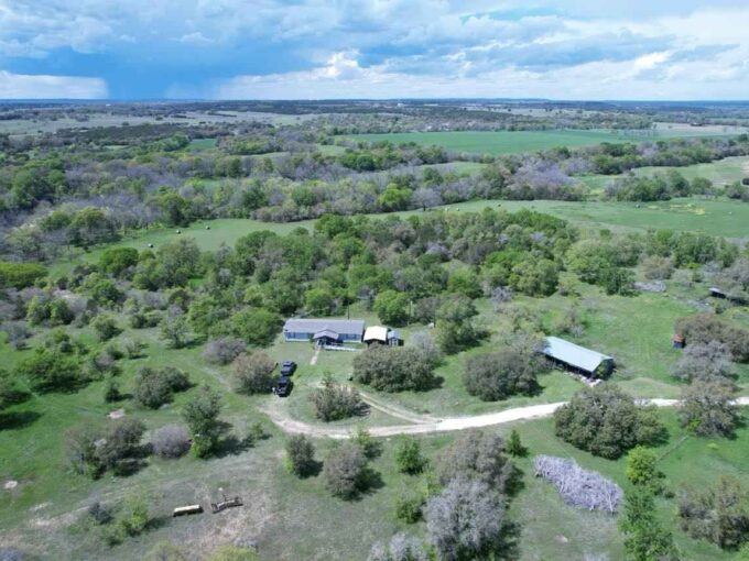 Aerial view of a rural property with a house, barn, scattered vehicles, and trees, surrounded by expansive green fields and distant hills under a partly cloudy sky.