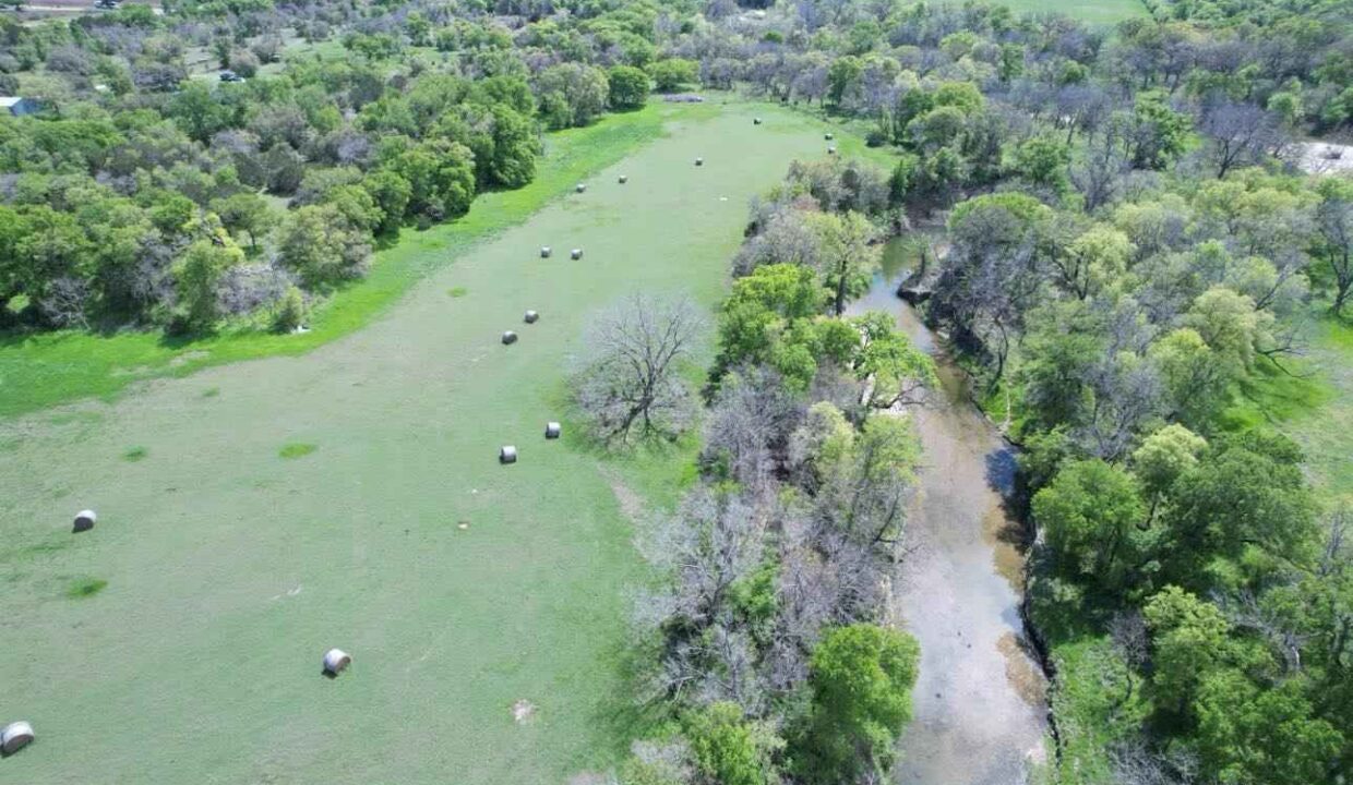 Aerial view of a green pasture with scattered hay bales, bordered by a winding creek and dense trees under a clear sky, with fields and forests in the distance.