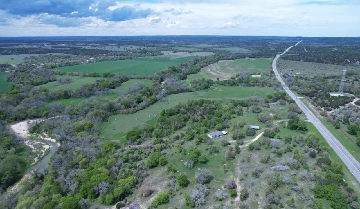Aerial view of a rural landscape with green fields, scattered trees, a winding road, and a few small buildings under a partly cloudy sky.