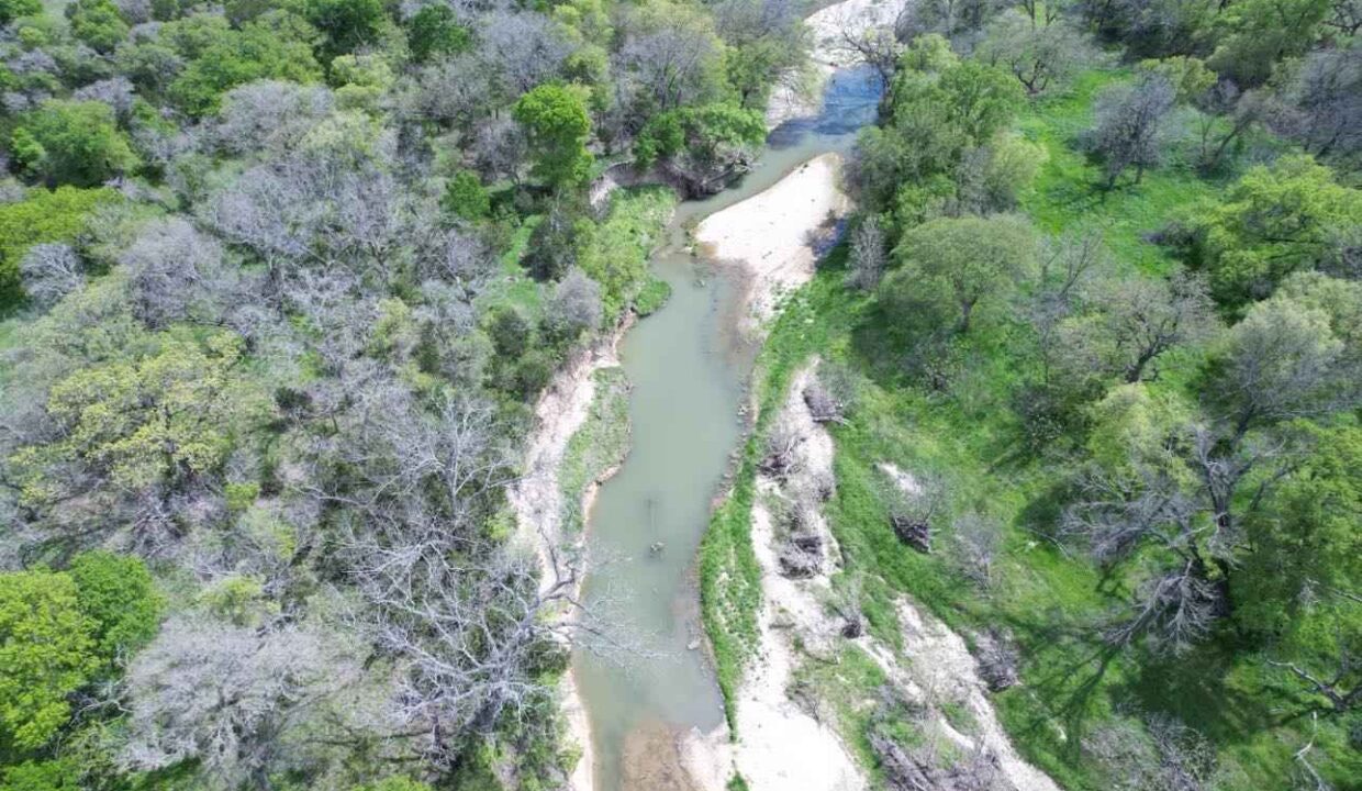 Aerial view of a narrow, winding river bordered by sandy banks and dense green trees with patches of dry foliage in a natural landscape.
