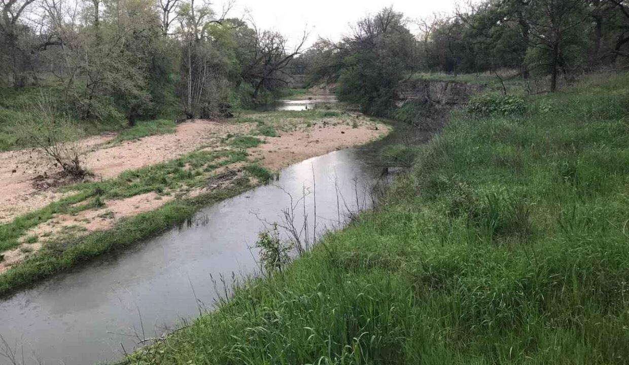A narrow creek winds through a grassy landscape with patches of sand and scattered trees, some with sparse leaves, under an overcast sky.