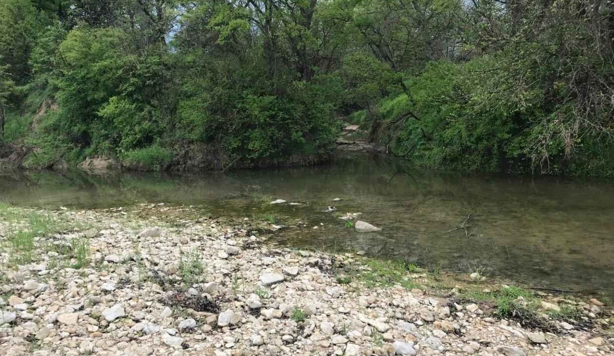 A shallow creek with clear water flows beside a rocky, pebbled shore, surrounded by dense green trees and bushes under a cloudy sky.