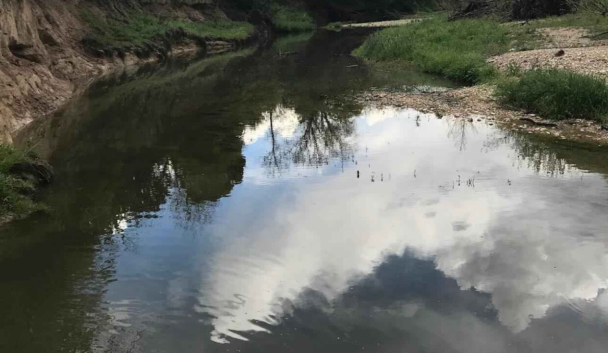 A calm river curves through a lush, green landscape with trees on both sides. The sky, filled with clouds, is reflected clearly in the water, creating a serene and peaceful scene.