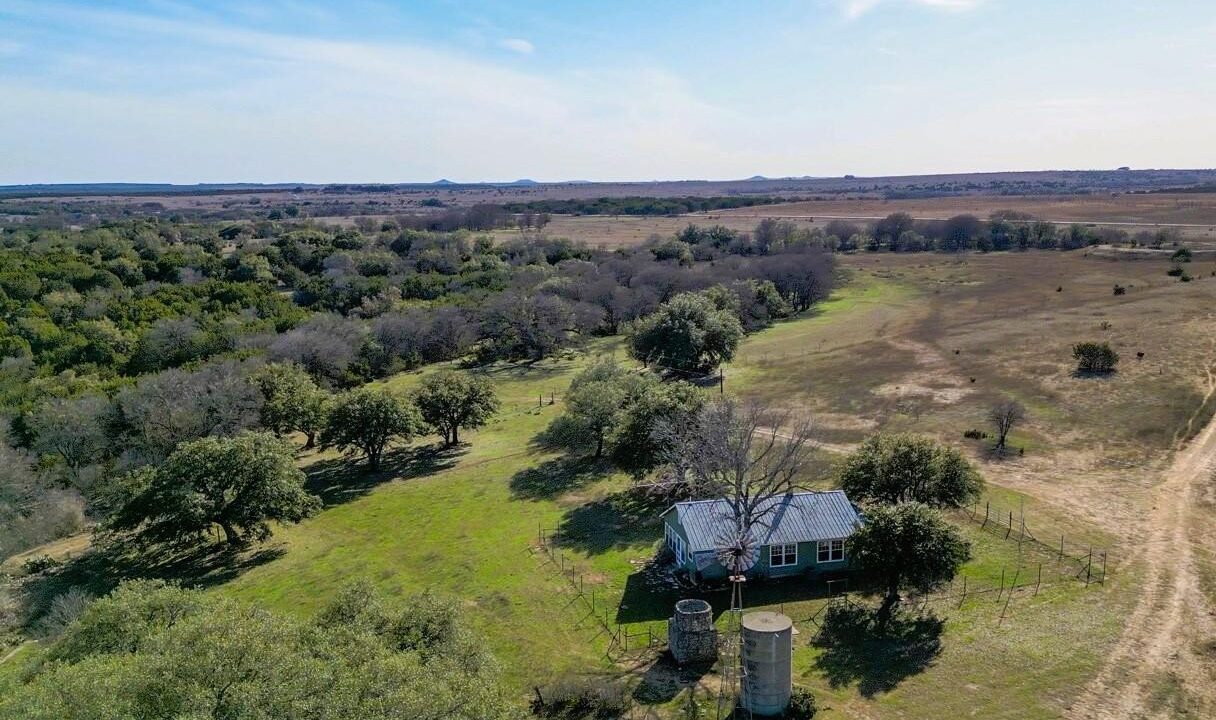 Aerial view of a rural landscape with a small house, two silos, scattered trees, and open fields under a blue sky. The land extends into the distance, with a mix of green and brown vegetation.
