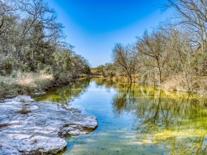 A clear, shallow stream flows between leafless trees and brush under a bright blue sky. Rocky banks and patches of grass line the water’s edge, and reflections of the trees are visible on the water’s surface.