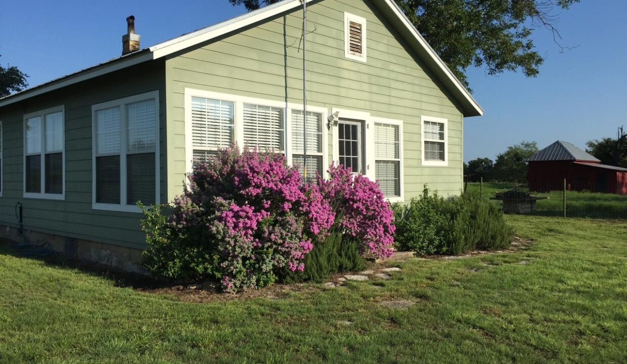 A small green house with white trim sits on a grassy lawn, bright pink and purple flowers blooming by the front door. A red barn stands in the background under a clear blue sky.